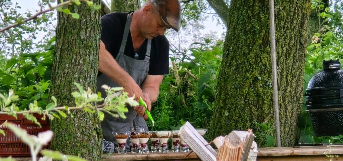Wijngaardenier Sico de Moel in schort dresseert dessertglaasjes op een houten tafel tussen de bomen, met keramische barbecue op de achtergrond.