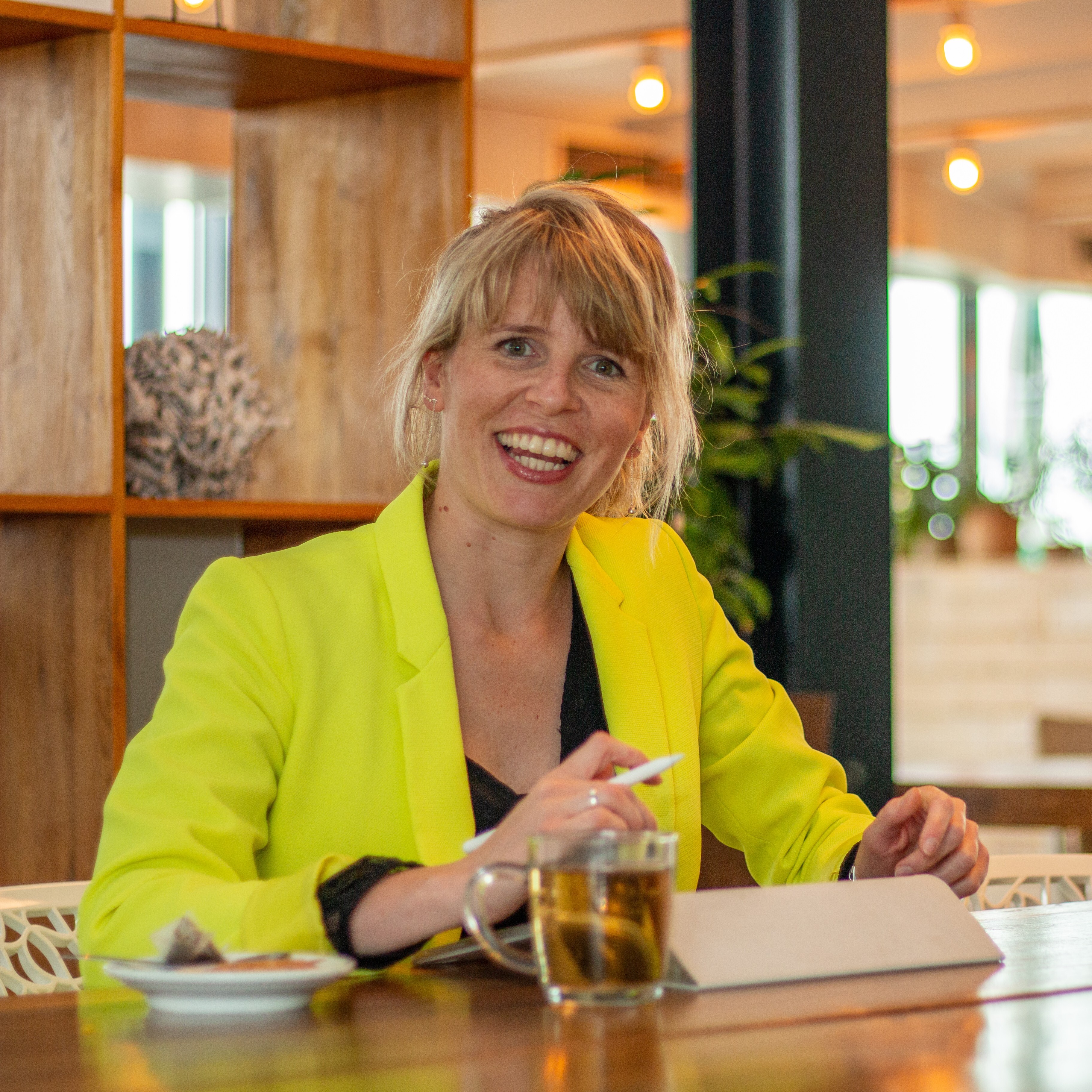 Foto Nanet Hak aan tafel met iPad voor zich, lachend naar de camera