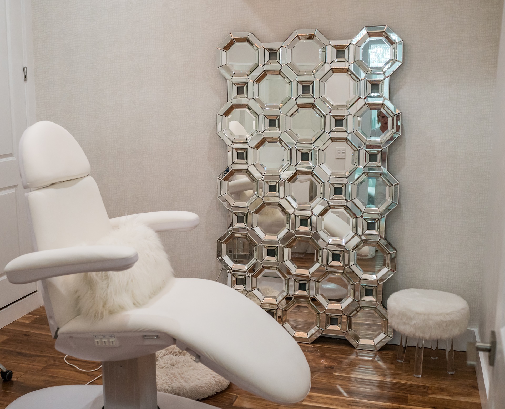 Behandelkamer, witte behandelstoel staat middenin de behandelkamer. Aan de wand hangt een decoratieve spiegel en een witte beklede kruk met witte vacht.