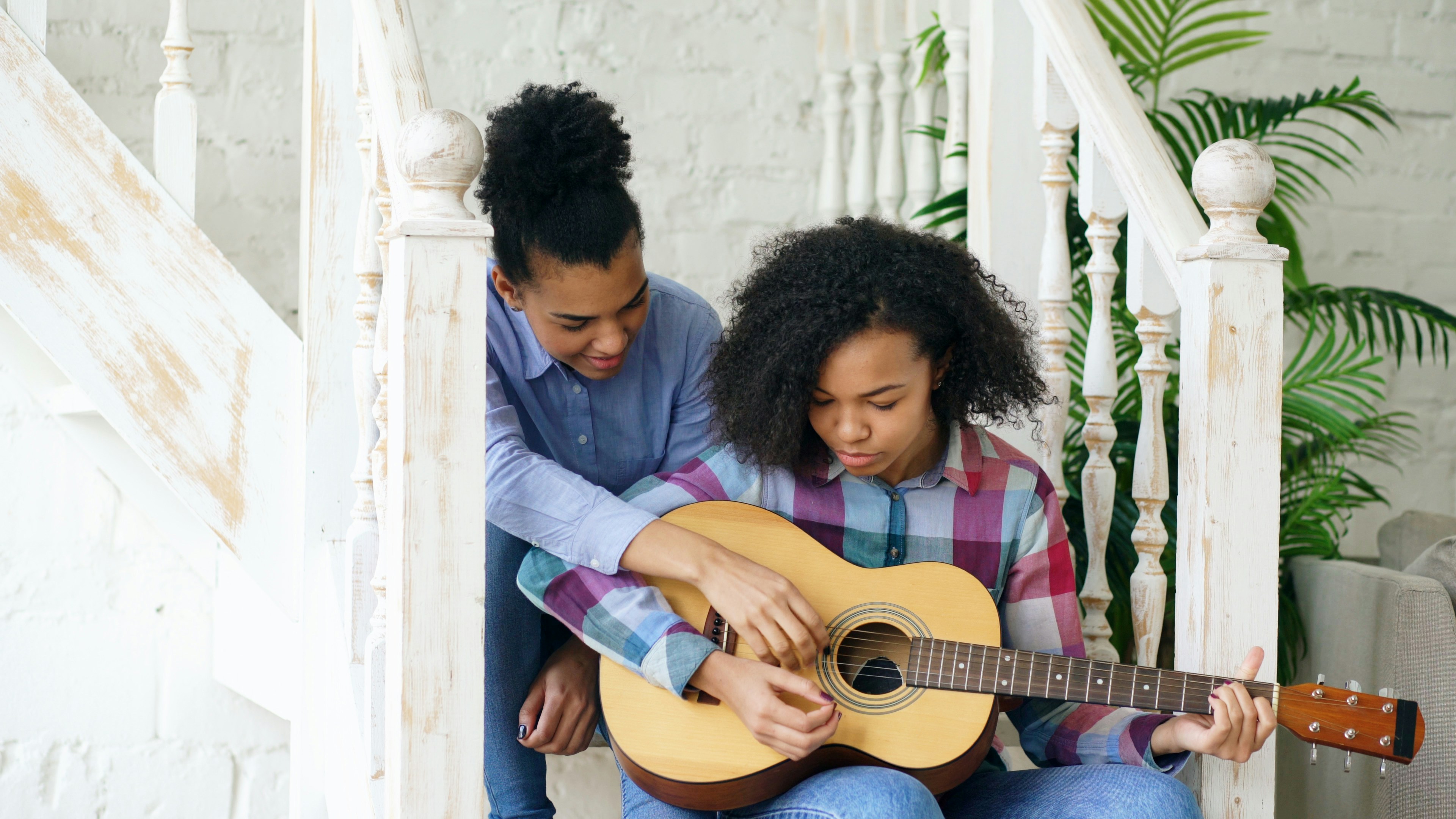Mixed race young woman sitting on stairs teaching her teenage sister to play acoustic guitar