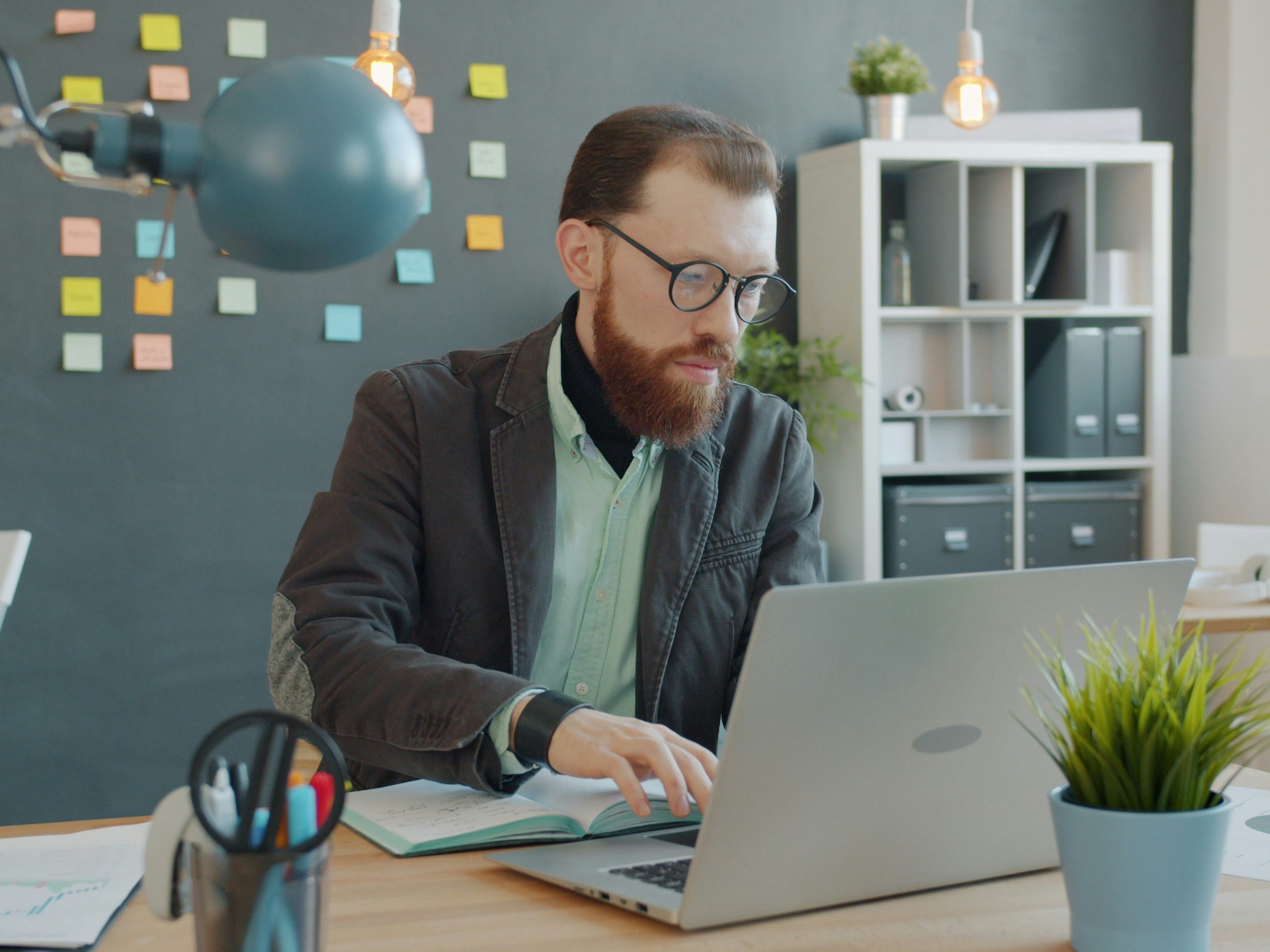 Serious man is using laptop typing then writing in notebook sitting at table in office concentrated on job. Modern technology, young people and occupation concept.