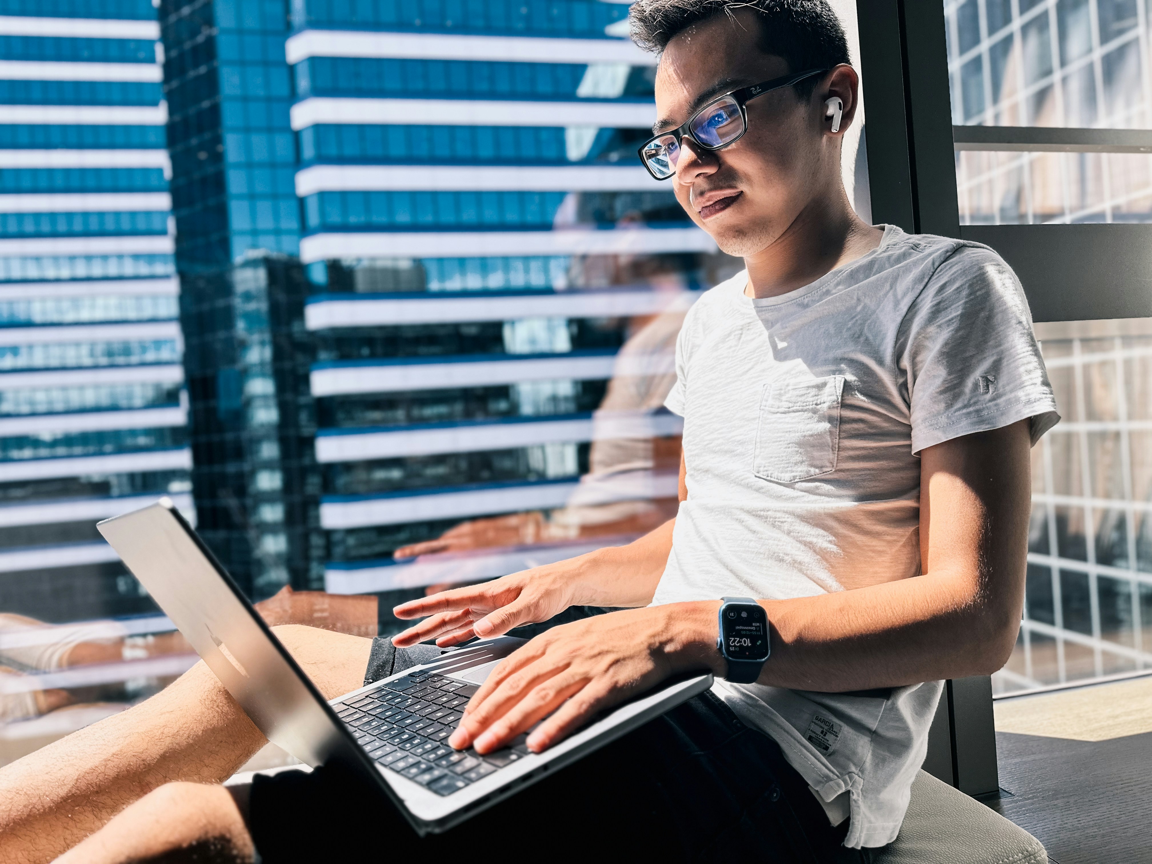 A young digital native is sitting on a window sill, wearing glasses and earbuds. He has his legs crossed and is using a laptop computer that rests on his lap. The sun shines through the window behind him, illuminating his face with its warm light. In back