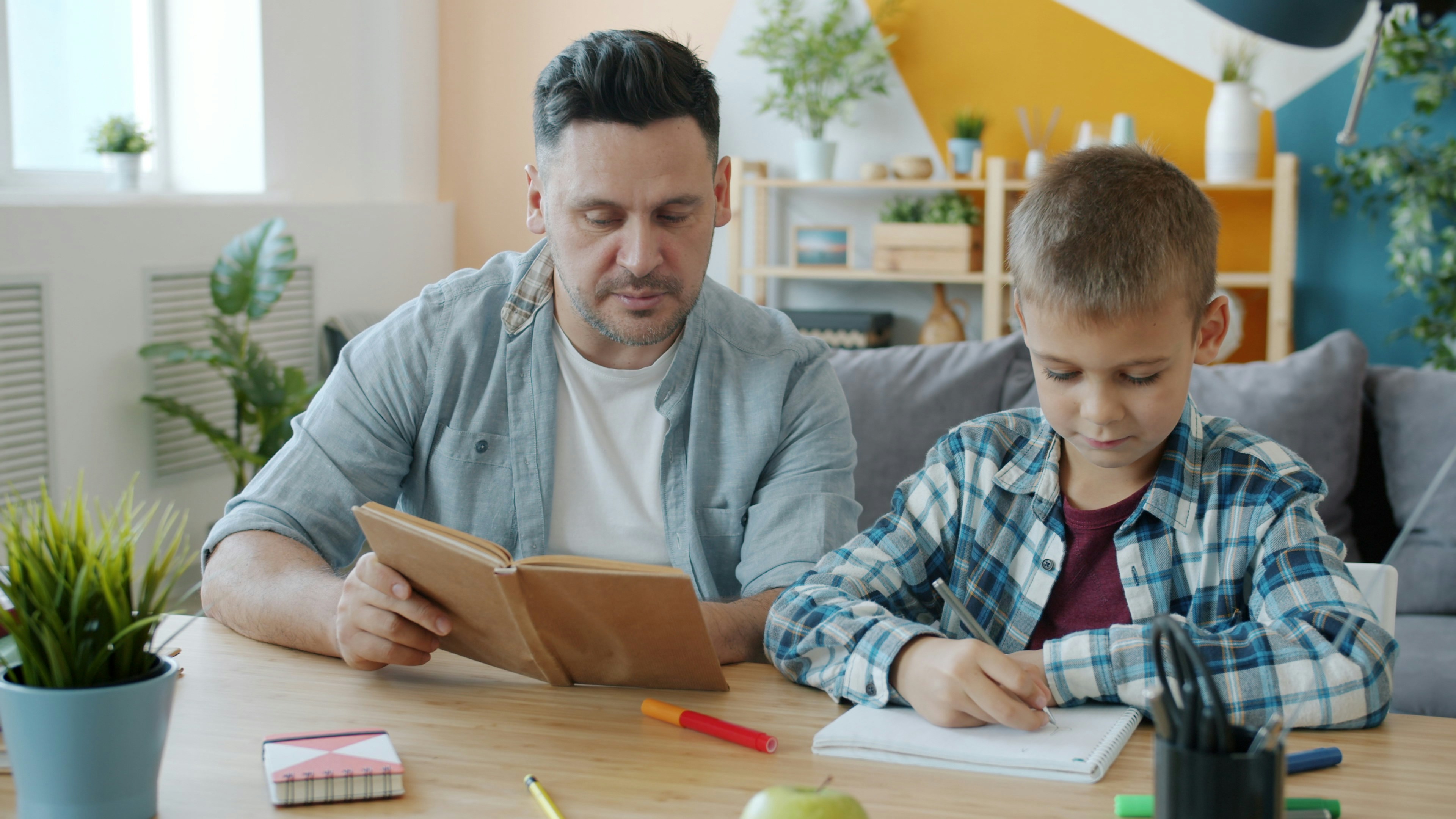 Father and son are drawing pictures and reading books having fun indoors at home, cozy apartment is visible in background. Literature and arts concept.