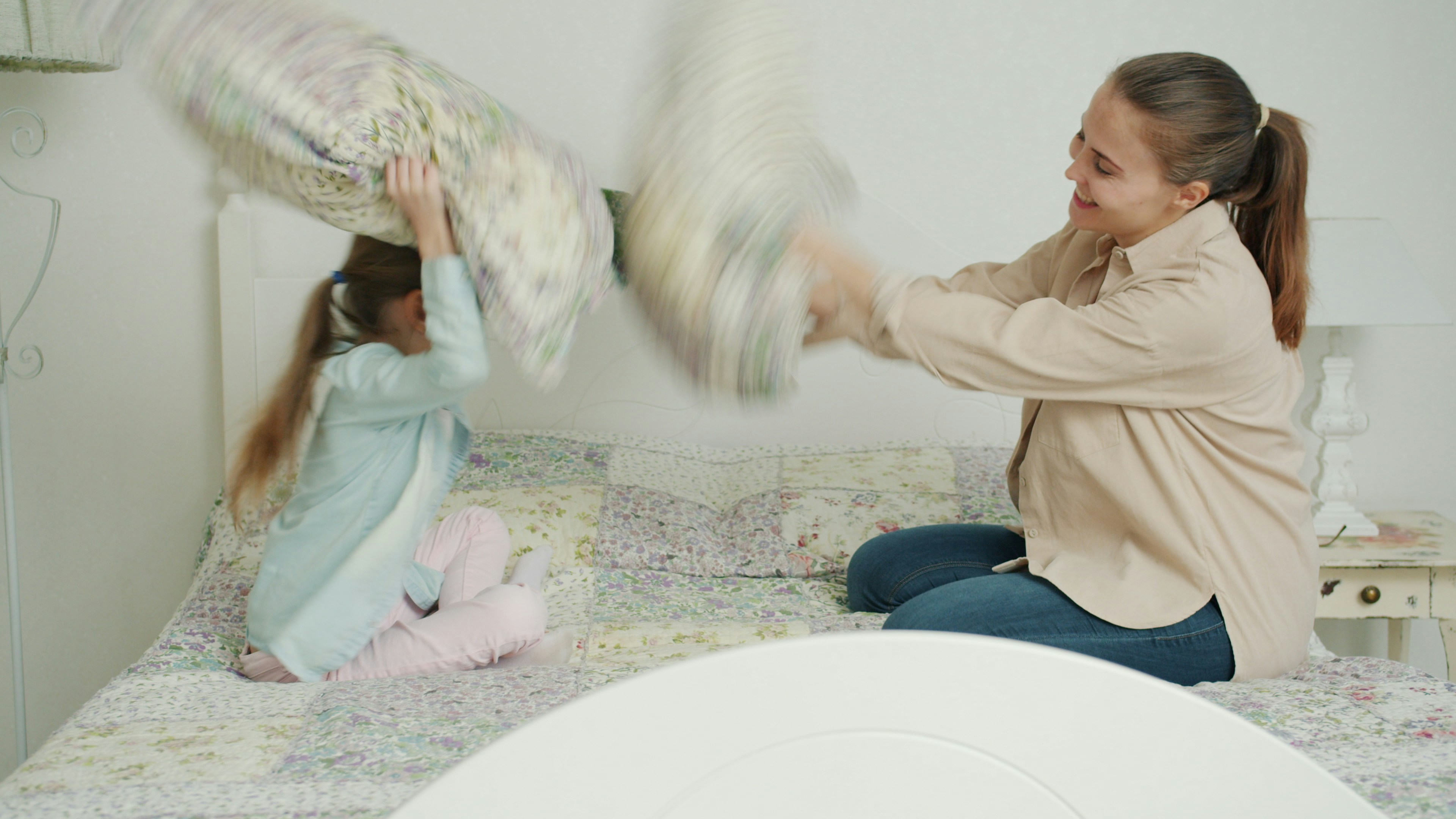 Little girl and loving mother are fighting pillows laughing relaxing in bed at home together, woman is playing with adorable child. Happiness and motherhood concept.