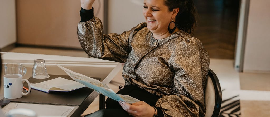 Vrouw met opgeheven hand tijdens training, glimlachend met een planning in de hand, symbool voor overzicht, zelfvertrouwen en eigenaarschap over je tijd