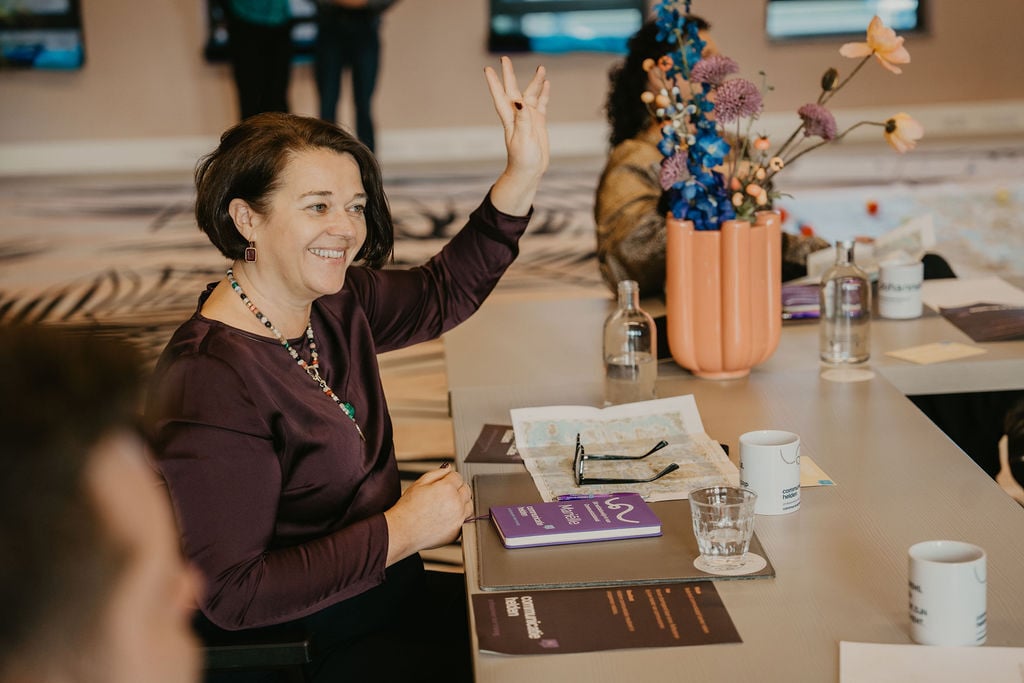 Vrouw steekt haar hand op tijdens een interactieve training over schrijven met AI, omringd door notities, boeken en frisse energie.