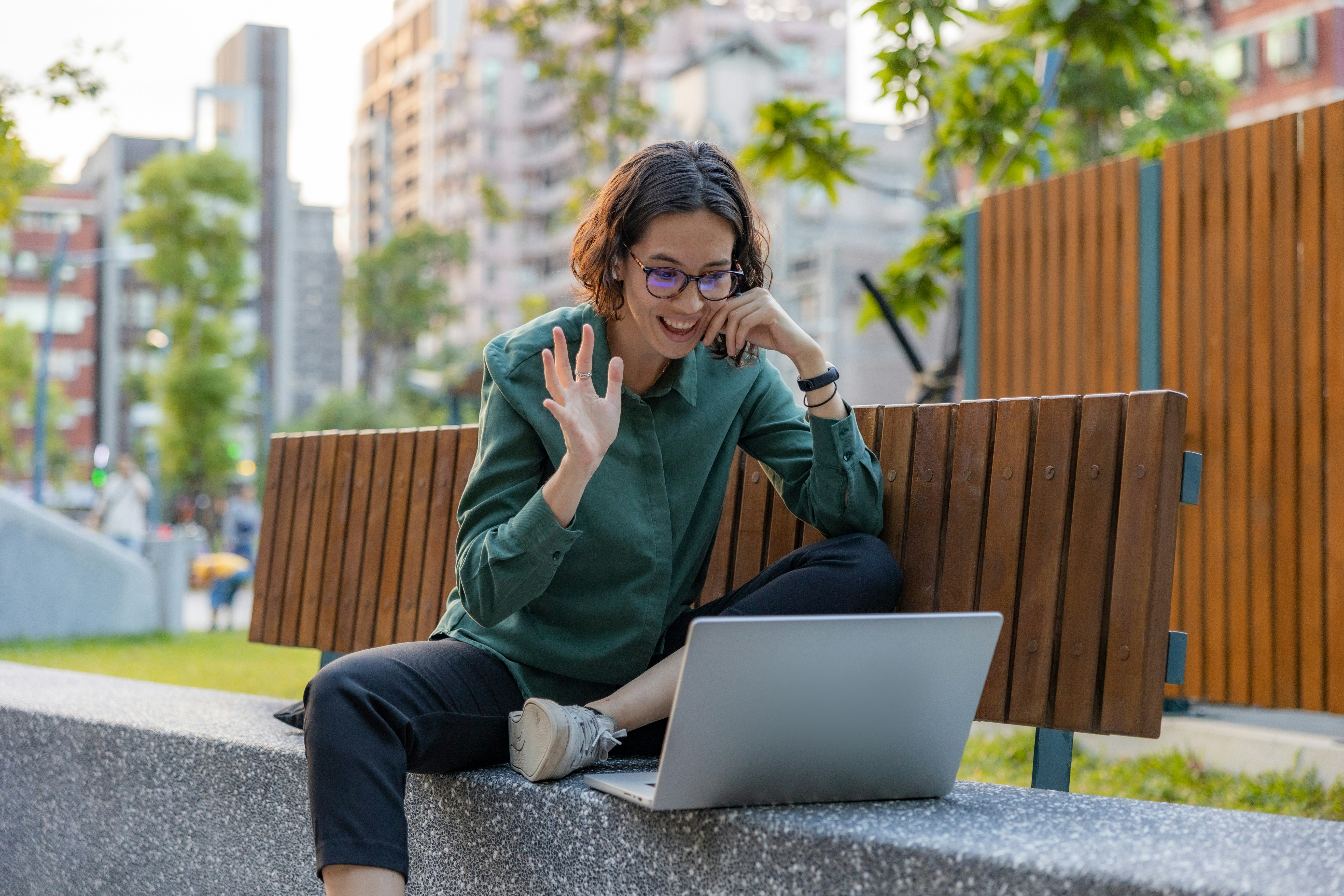 Smiling young woman waving hello and doing remote work video call at park bench